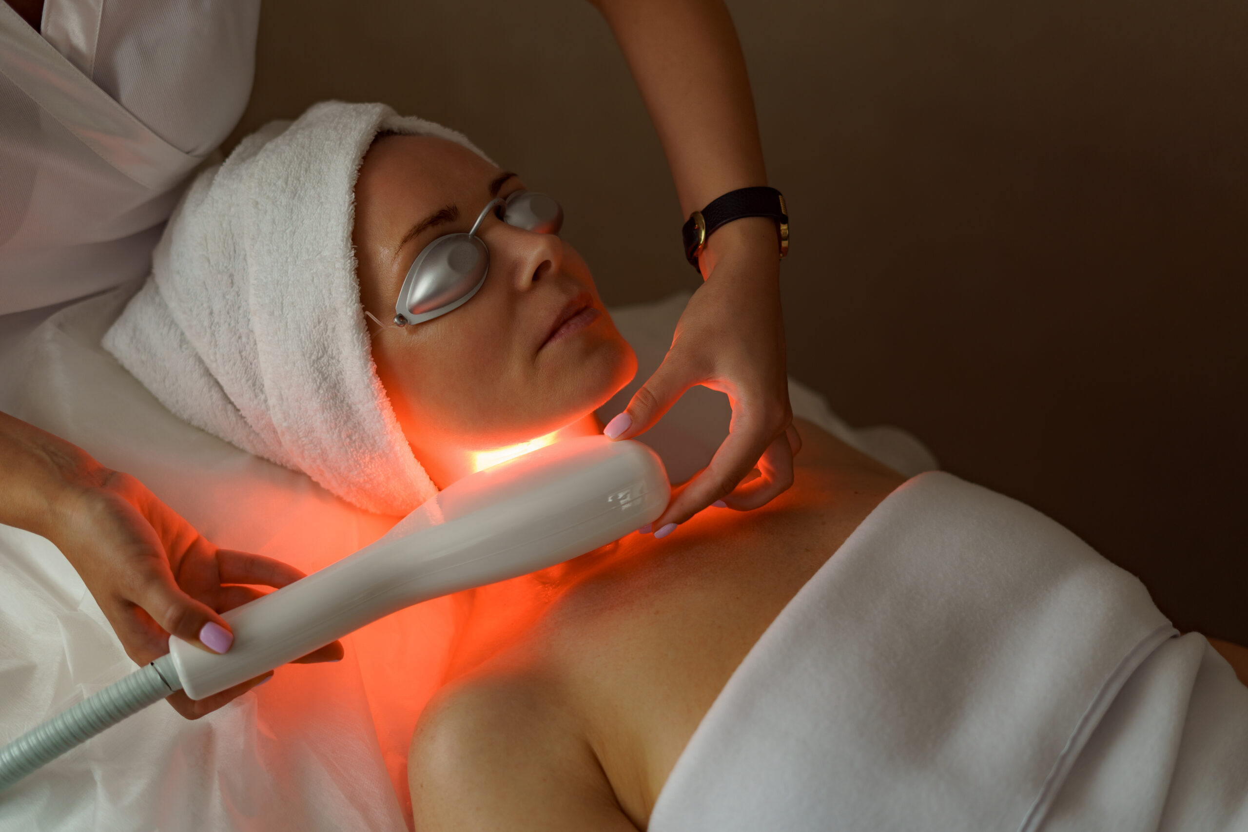View of doctor cosmetologist doing anti aging procedure in cosmetology office. Satisfied woman in disposable hat lying on couch and relaxing. Working with Apparatus.